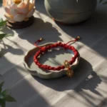 Red cord Buddhist bracelet, decorated with small golden charms, with a backdrop of Buddhist floral decorations, showcasing zen and fine craftsmanship.